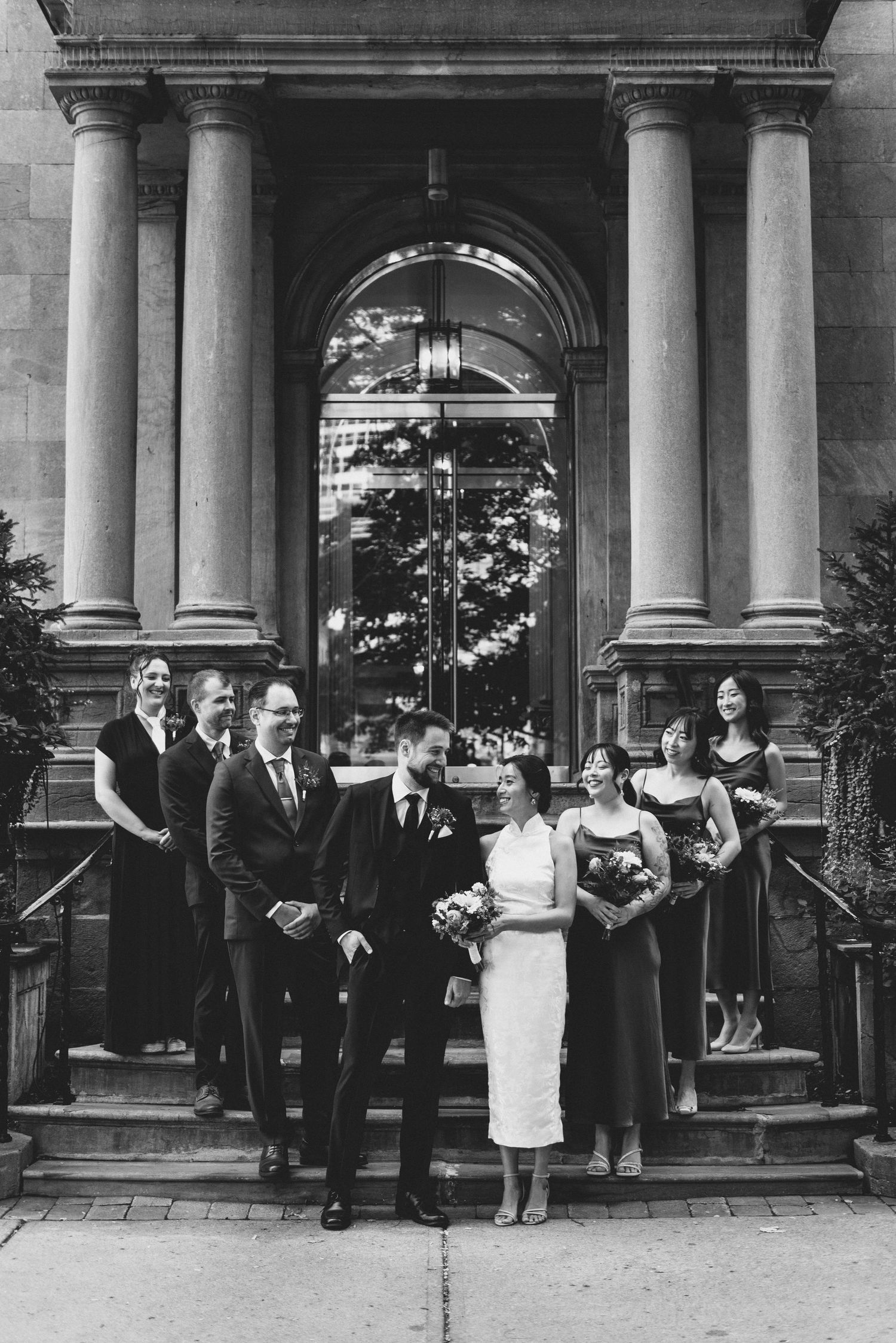 Black and white wedding party portrait standing on ornate stone steps with classical columns in the background.