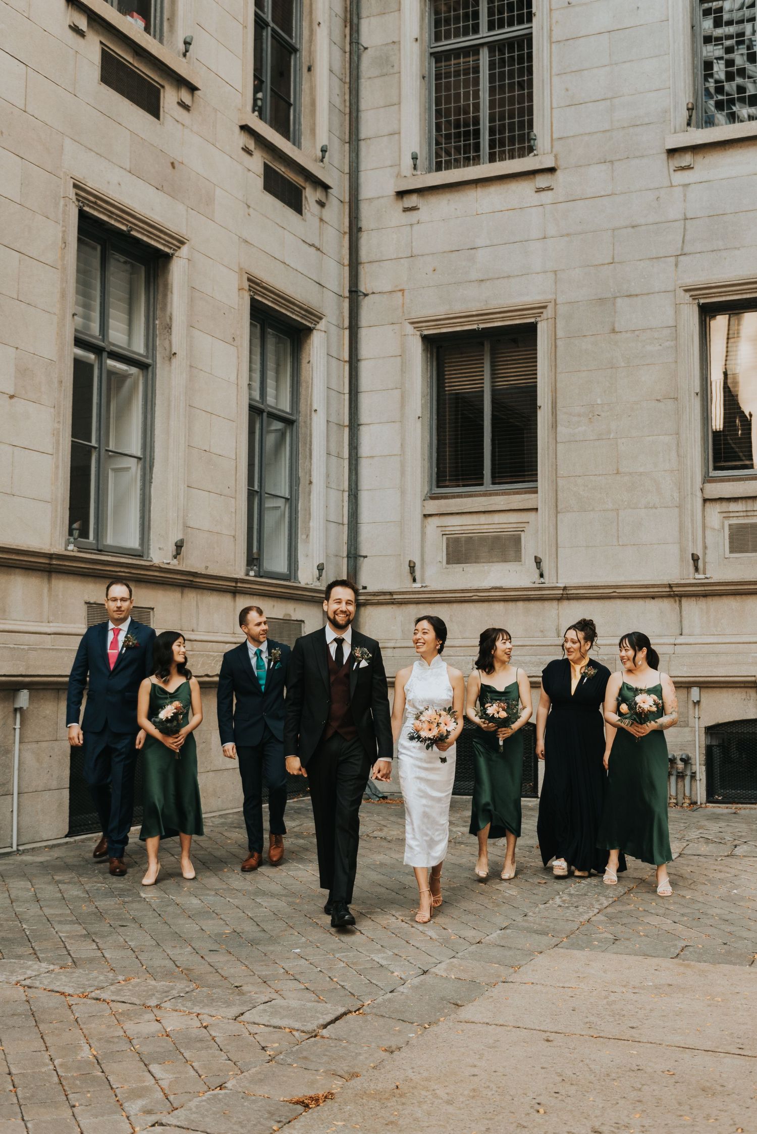 Wedding party in formal attire walking outside a grand stone building with tall windows.