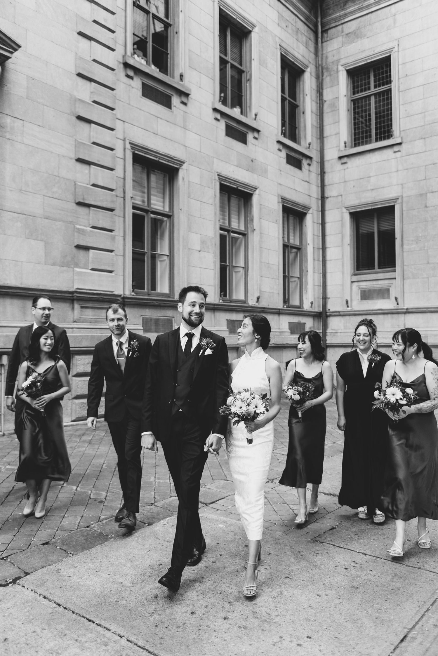 Wedding party walks together outside a classic stone building with elegant windows in black and white photography.
