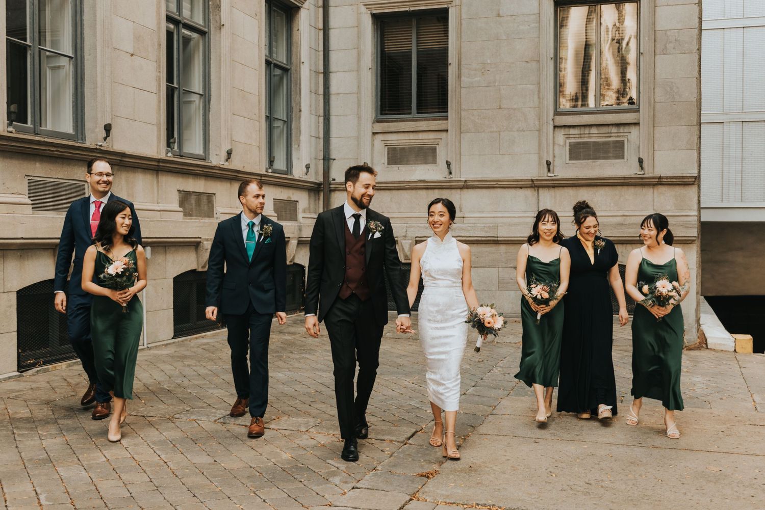 Wedding party in formal attire walking together outside a classic stone building with elegant architectural details.