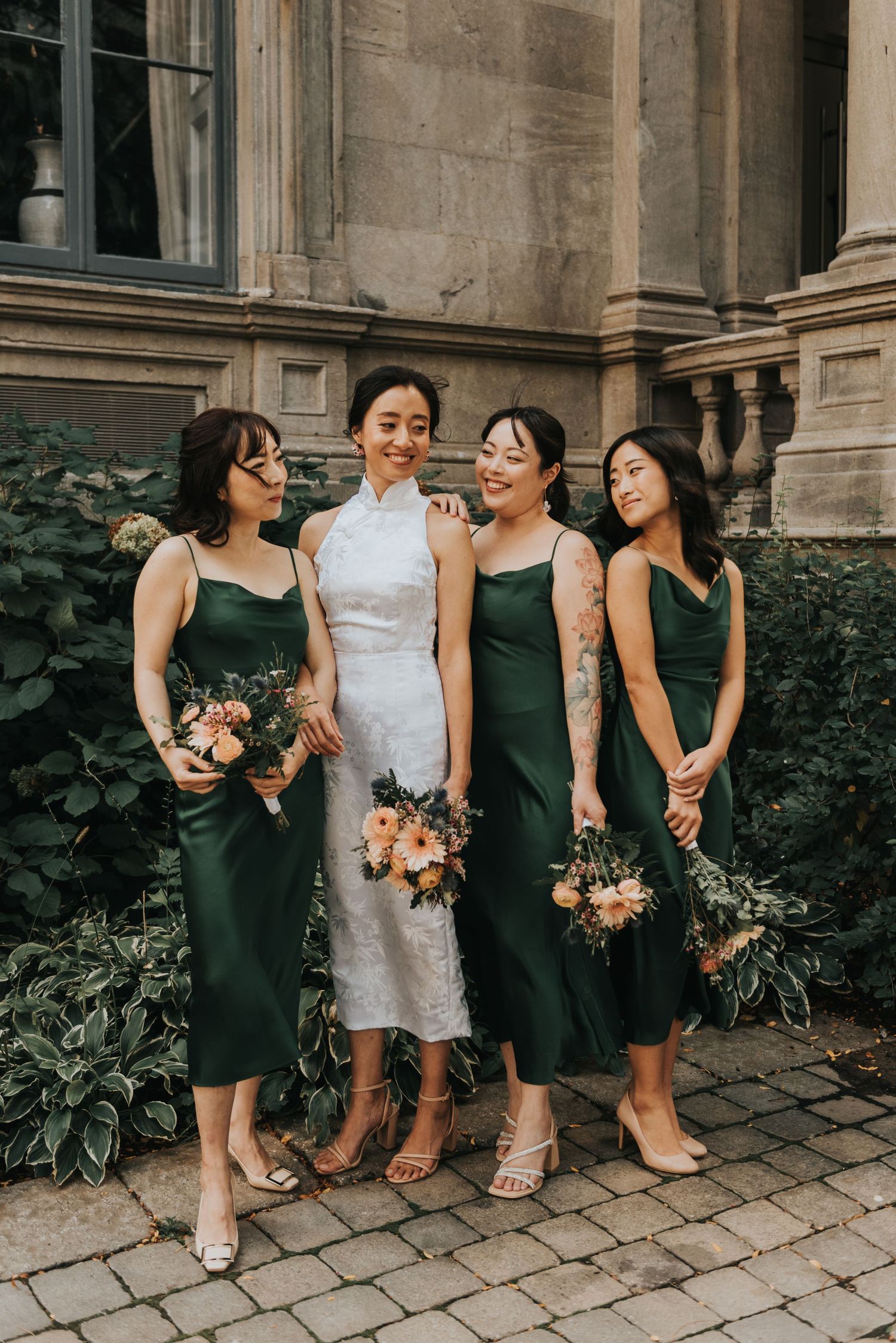 A bridal party poses outdoors in elegant dark green bridesmaid dresses holding floral bouquets against stone architecture.