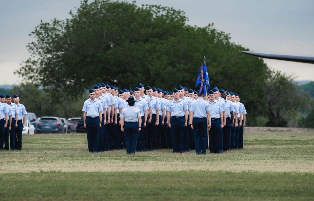 Military personnel in blue uniforms stand in formation on a grassy field with trees in the background.