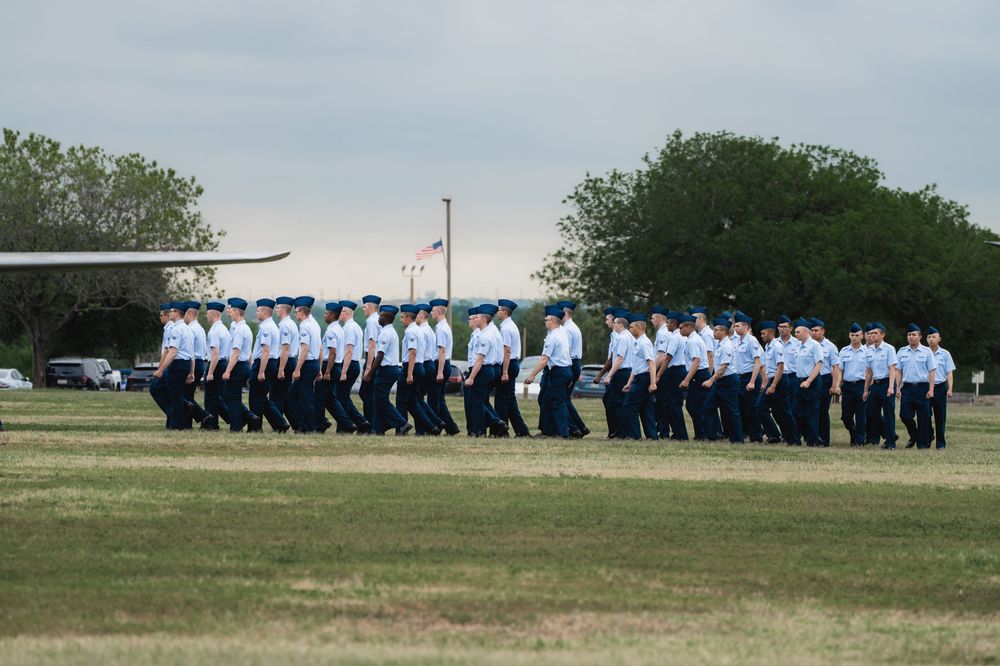 A group of people dressed in light blue and navy uniforms stand in formation on a grassy field during an outdoor ceremony.