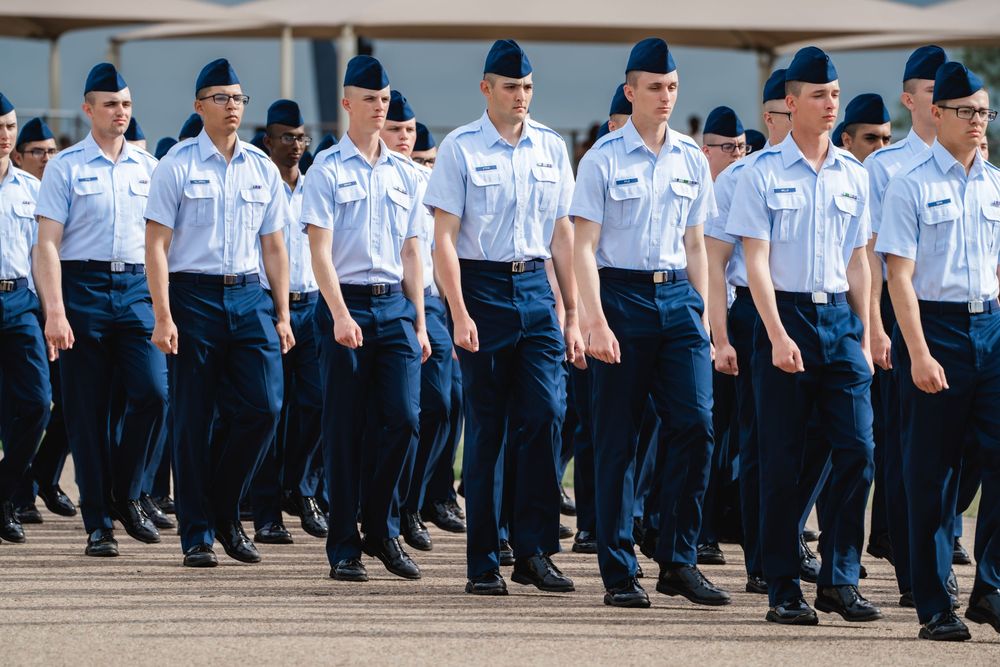 Military personnel in blue uniforms and caps march in formation on a paved surface.