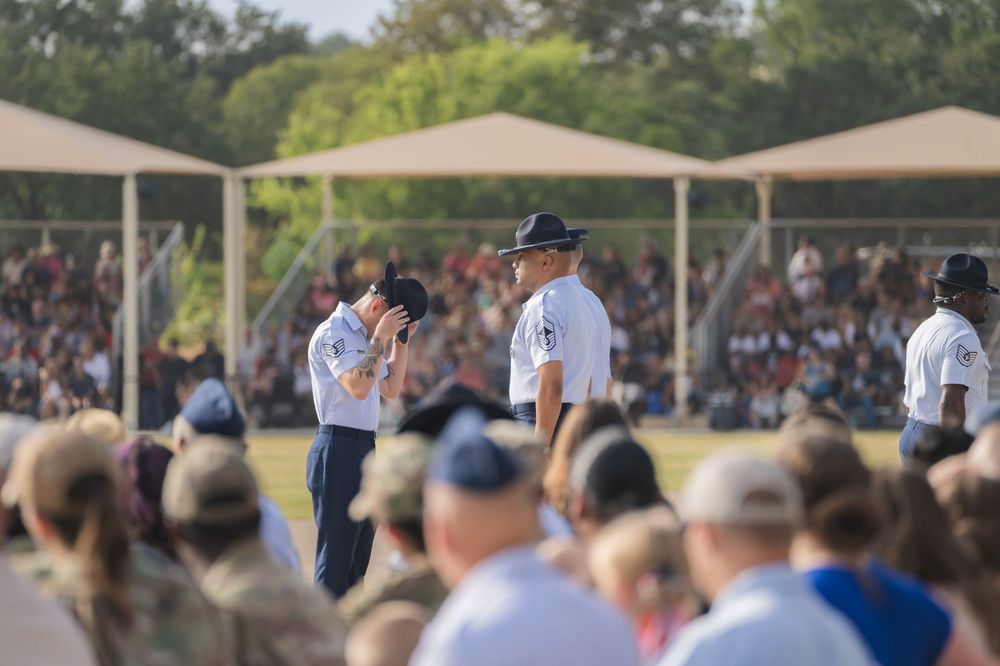 Military personnel stand on stage at an outdoor ceremony with crowd members seated under canopy tents in background.