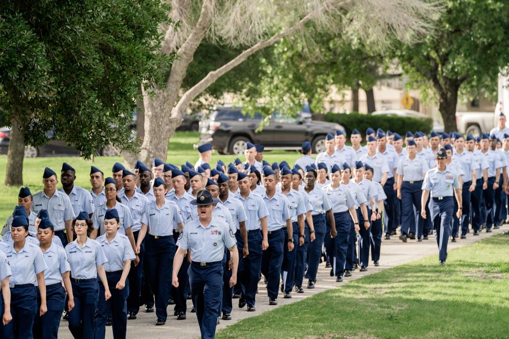 Group of Air Force cadets in blue uniforms marching in formation along tree-lined path on sunny day.