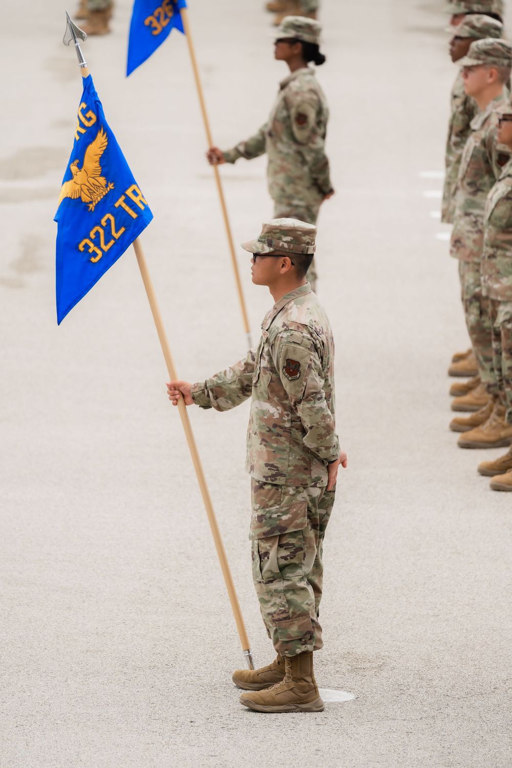 Military service members in camouflage uniforms stand at attention holding bright blue unit guidon flags during formation.