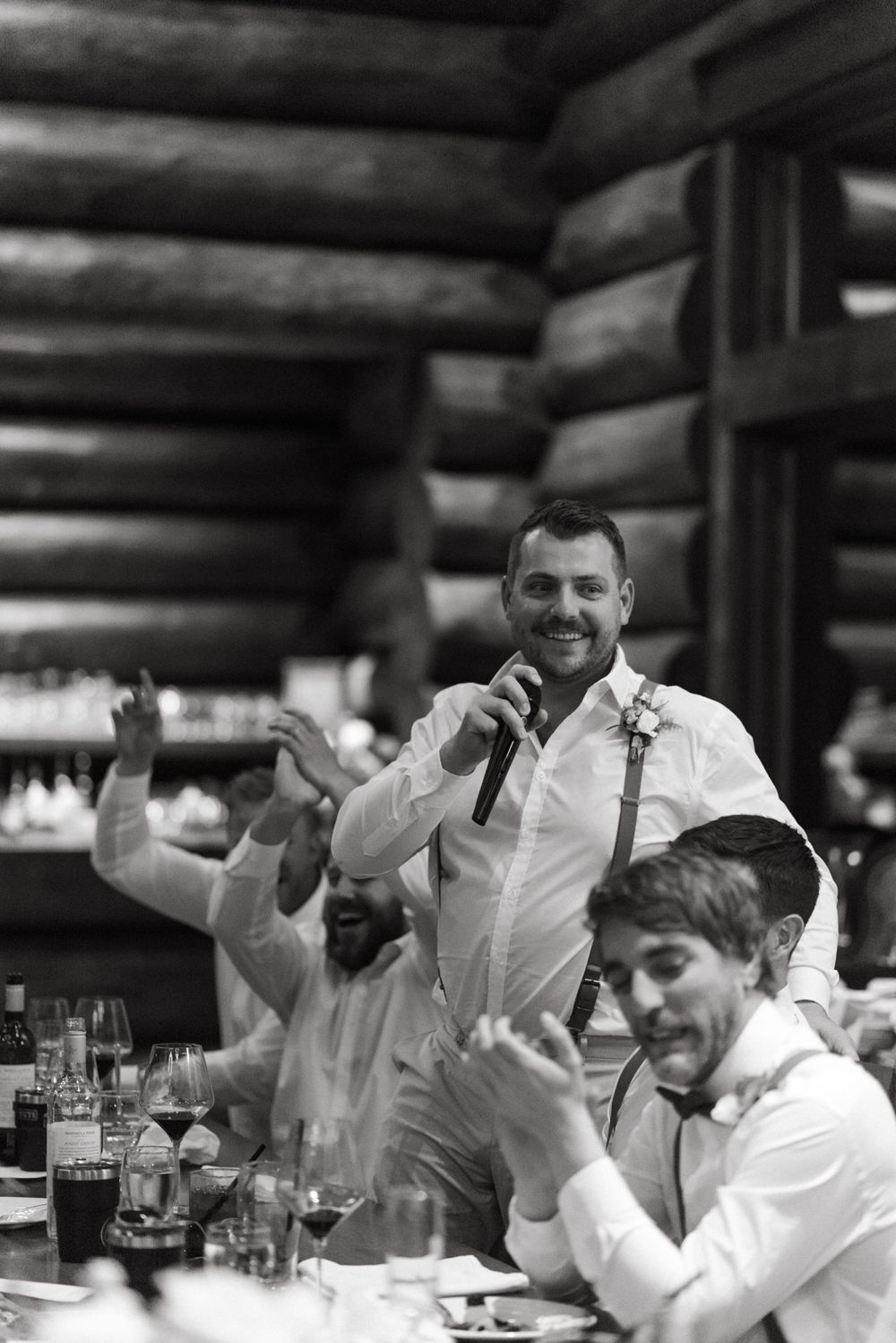 A black and white photo of guests at a wedding reception with a speech taking place in a rustic log cabin venue.