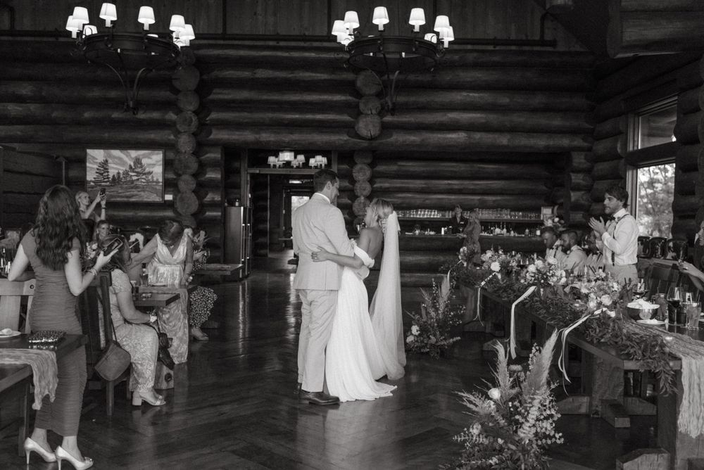 A couple shares their first dance in a rustic log cabin wedding venue with decorative lighting and guests watching.