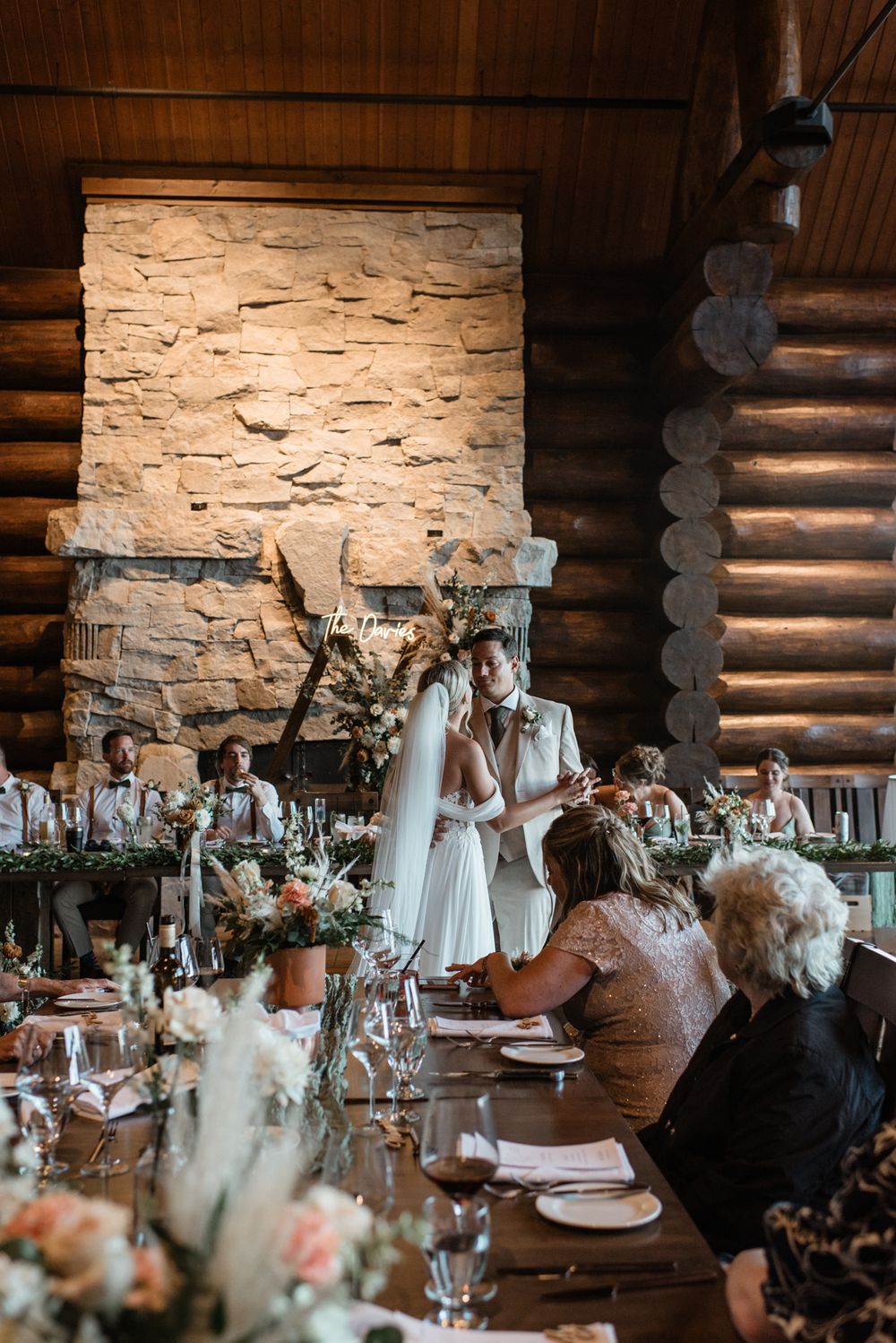 An elegant rustic wedding reception in a log cabin with stone fireplace and long tables decorated with floral arrangements.