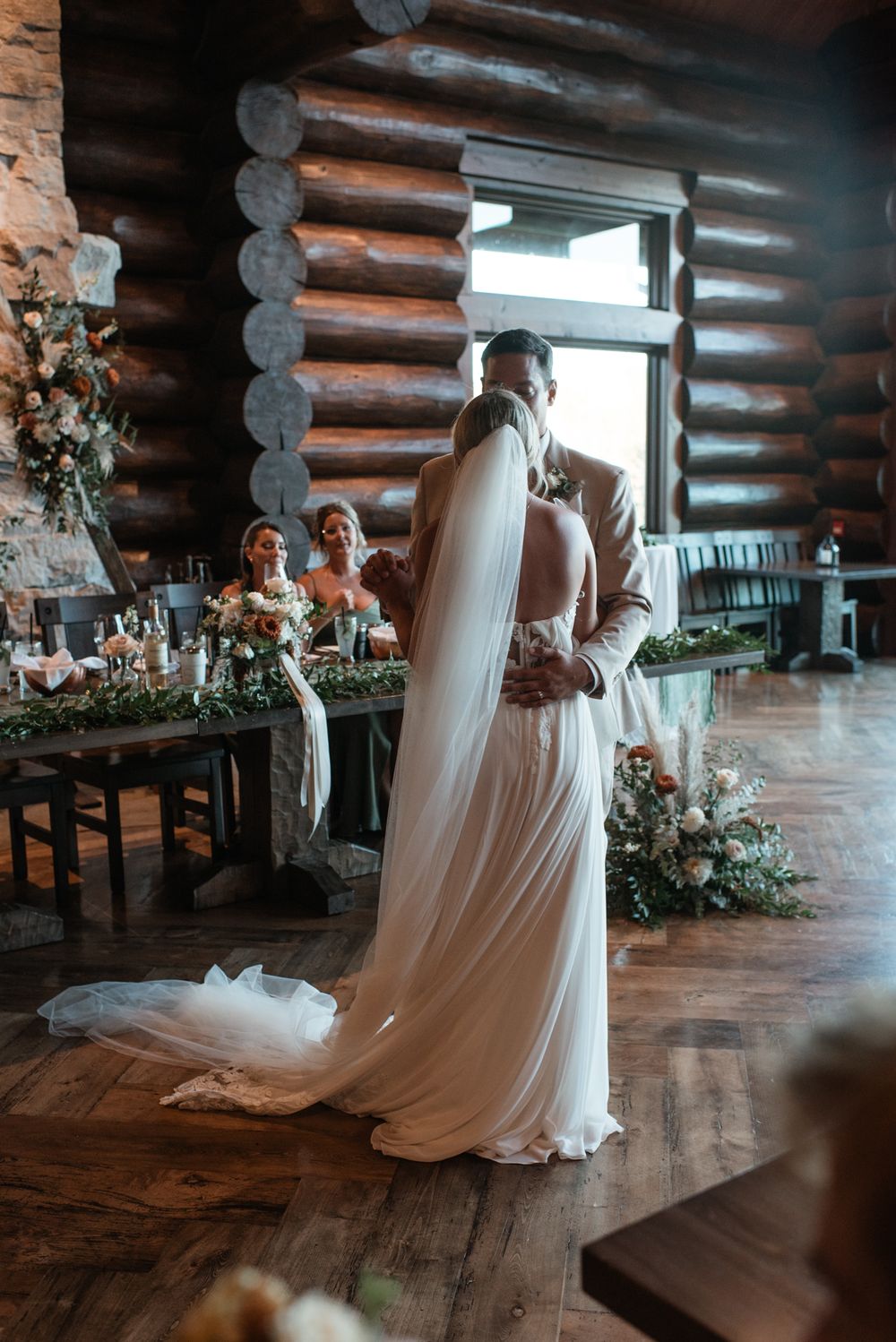 A wedding couple shares an intimate first dance moment inside a rustic log cabin venue with floral arrangements.