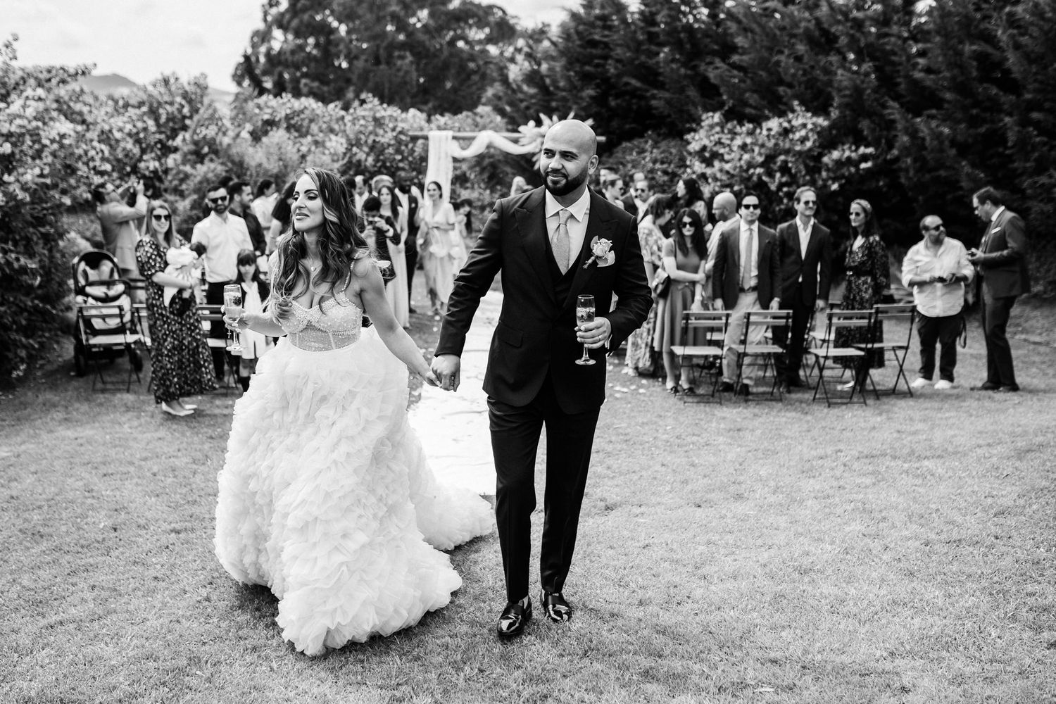 Black and white photo of a bride and groom walking hand in hand during an outdoor garden wedding ceremony.