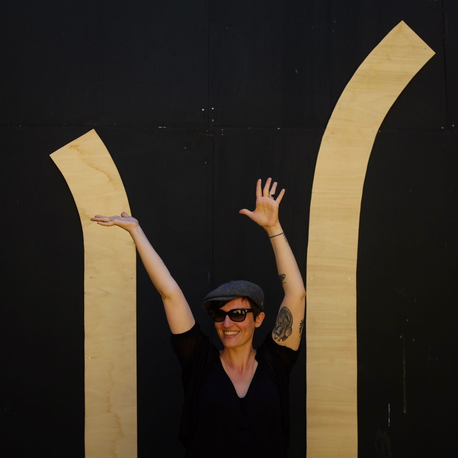 Person in black clothes playfully posing against dark wall with golden arch shapes.