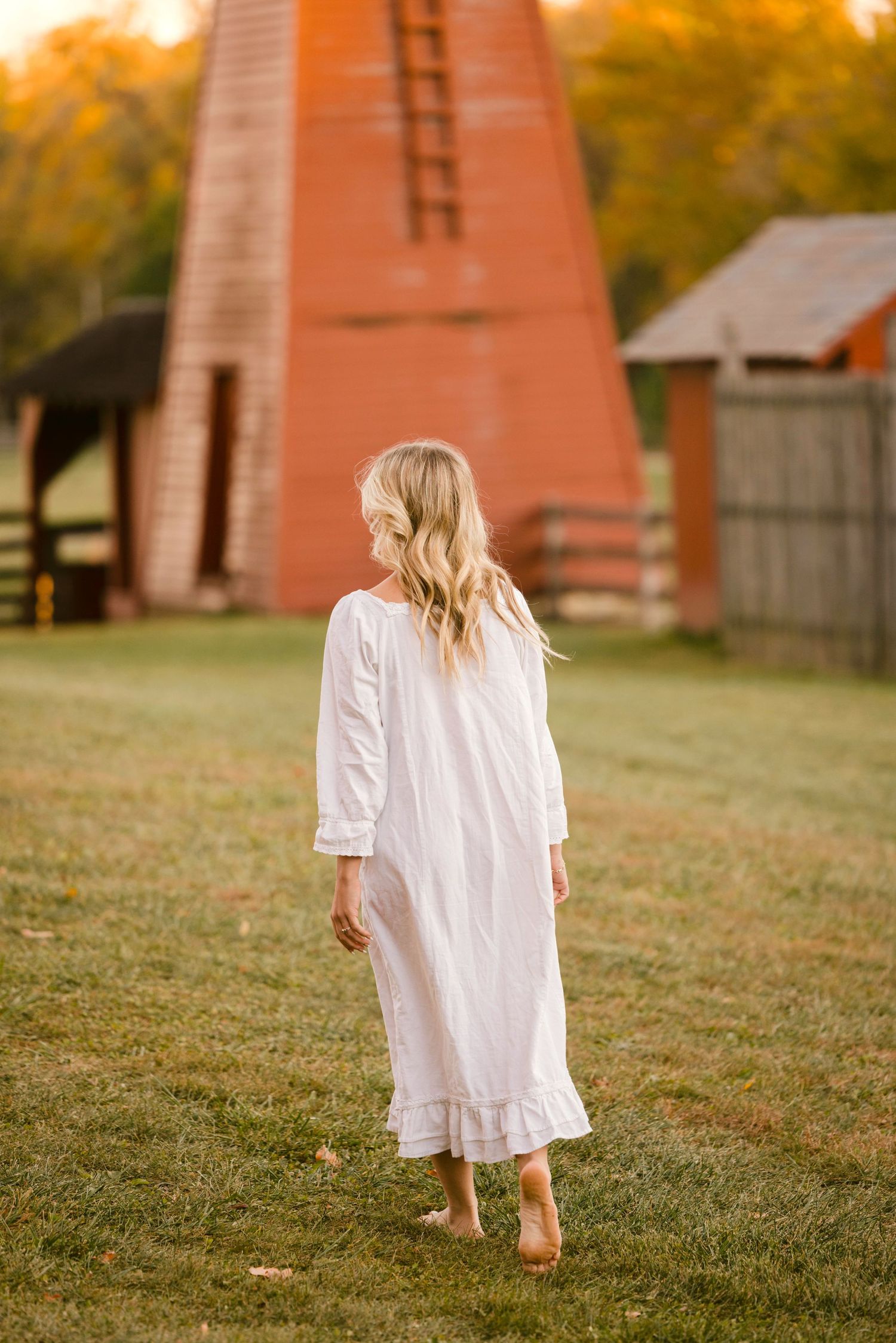 Person in white vintage nightgown stands on grass near orange windmill structure during autumn sunset.