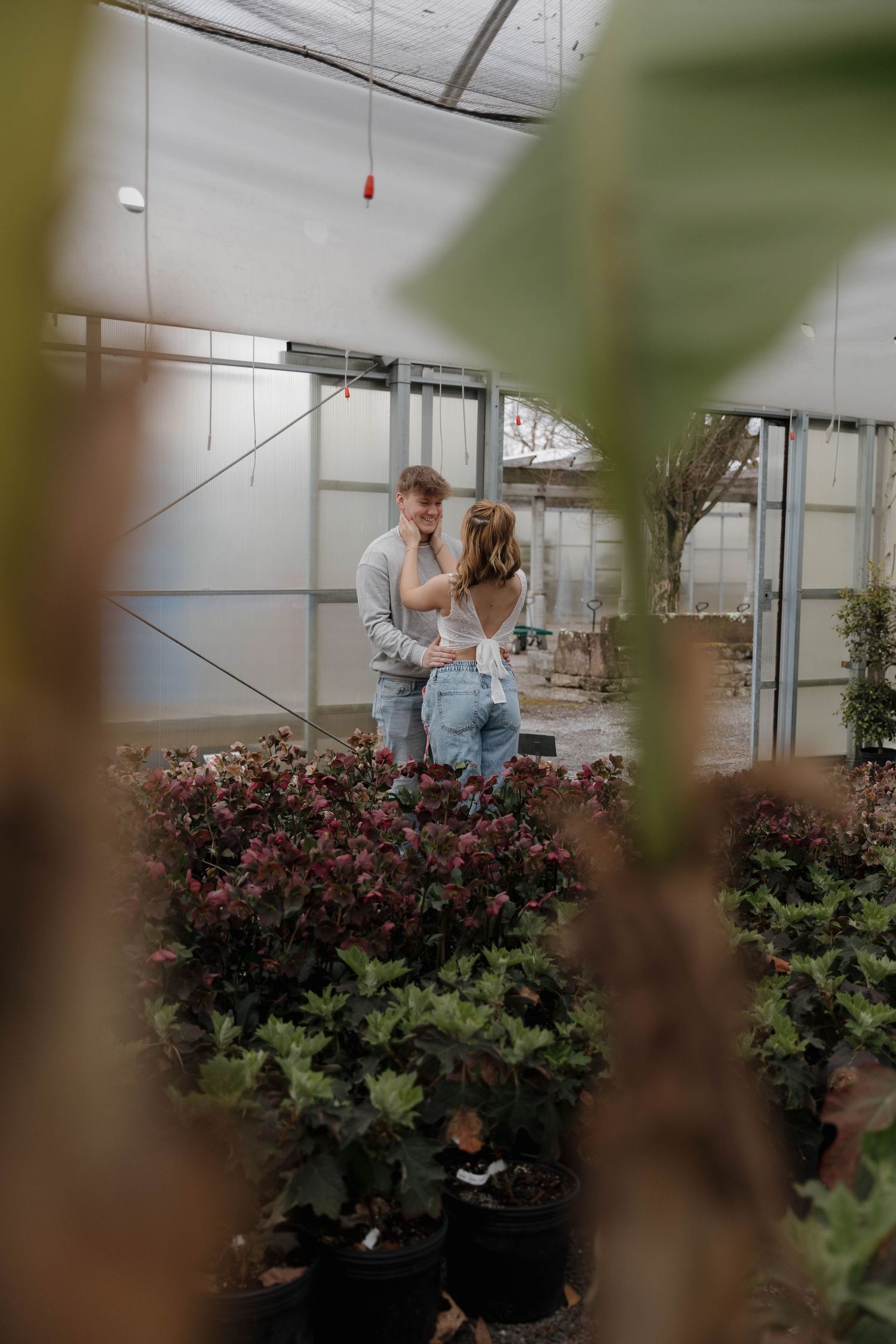 A couple embraces in a greenhouse surrounded by potted plants and flowering bushes.