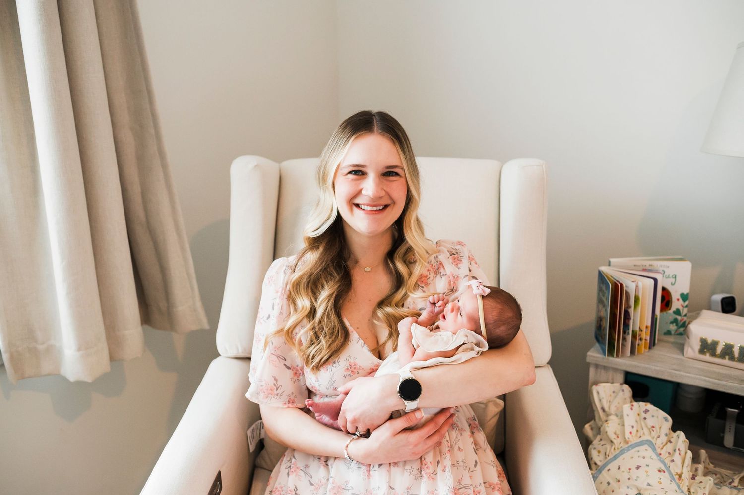 A tender moment in a nursery as a parent holds their sleeping newborn in a white armchair beside window light.
