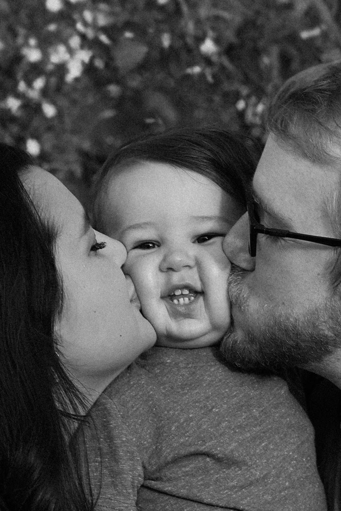 Black and white photo of loving parents kissing their smiling baby's cheeks in sweet family moment.