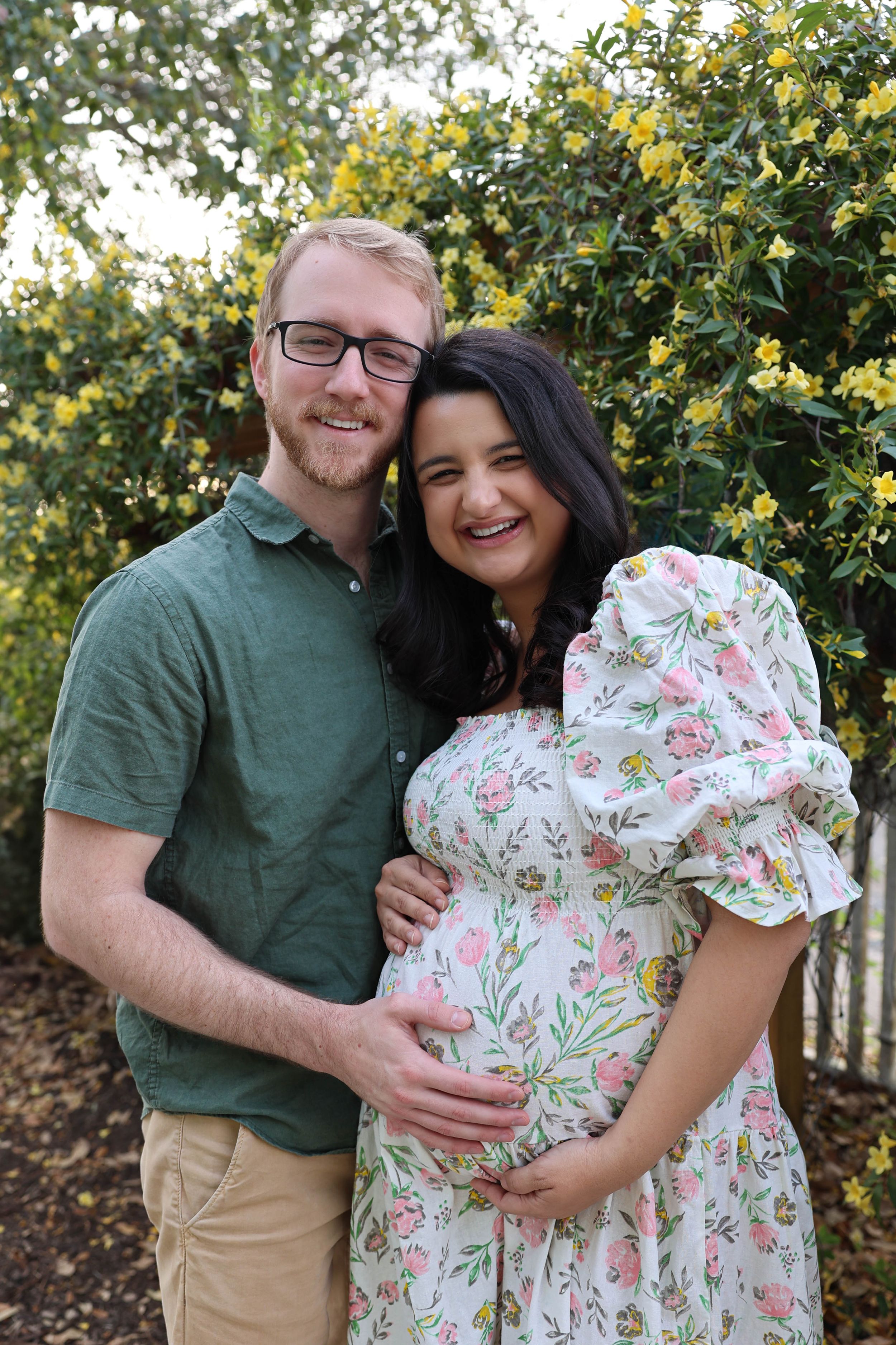 A couple poses together outdoors in front of yellow flowers, with the woman wearing a floral dress during pregnancy.