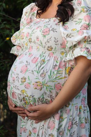 A pregnancy bump is cradled in a flowy white floral dress with pink and yellow blooms and smocked detailing.