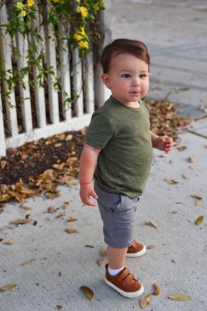 Small toddler wearing olive green t-shirt and grey shorts stands on sidewalk by white picket fence with yellow flowers.