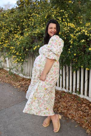 A pregnant woman in a floral dress poses next to a white picket fence with yellow flowers in the background.