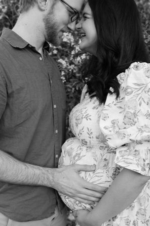 Black and white maternity photo showing a couple in casual clothing embracing outdoors, with hands resting on pregnant belly.