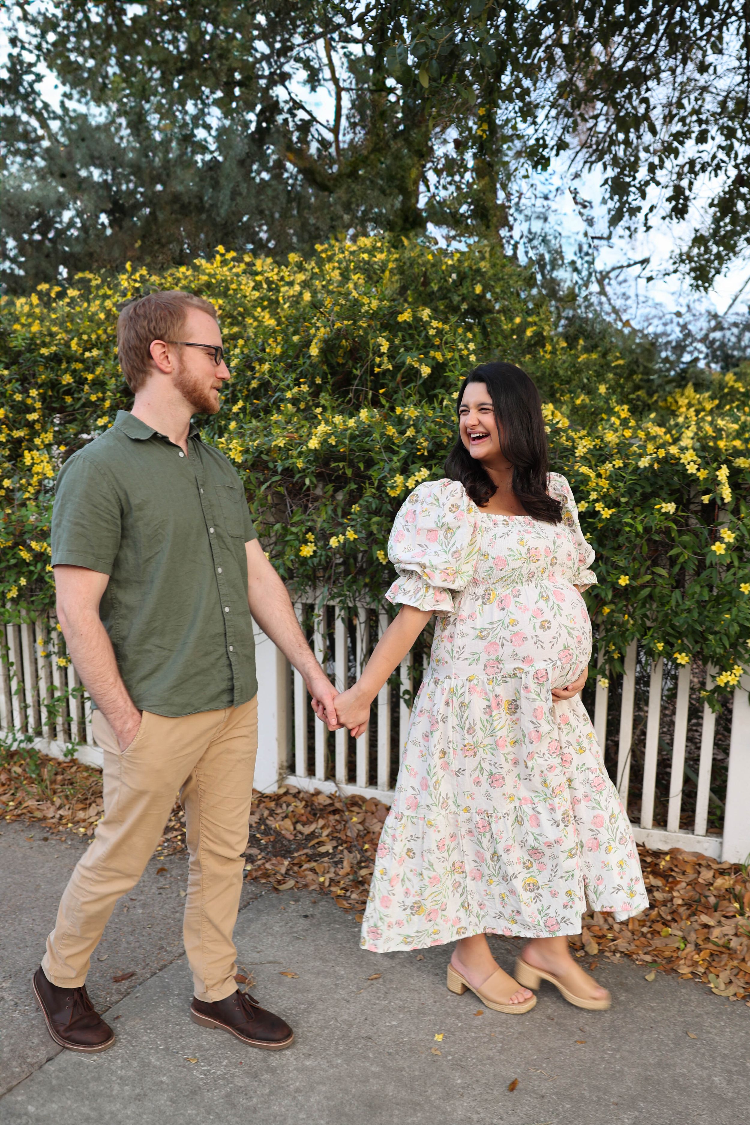 A couple holds hands while standing outdoors in front of a white picket fence and yellow flowering shrubs.