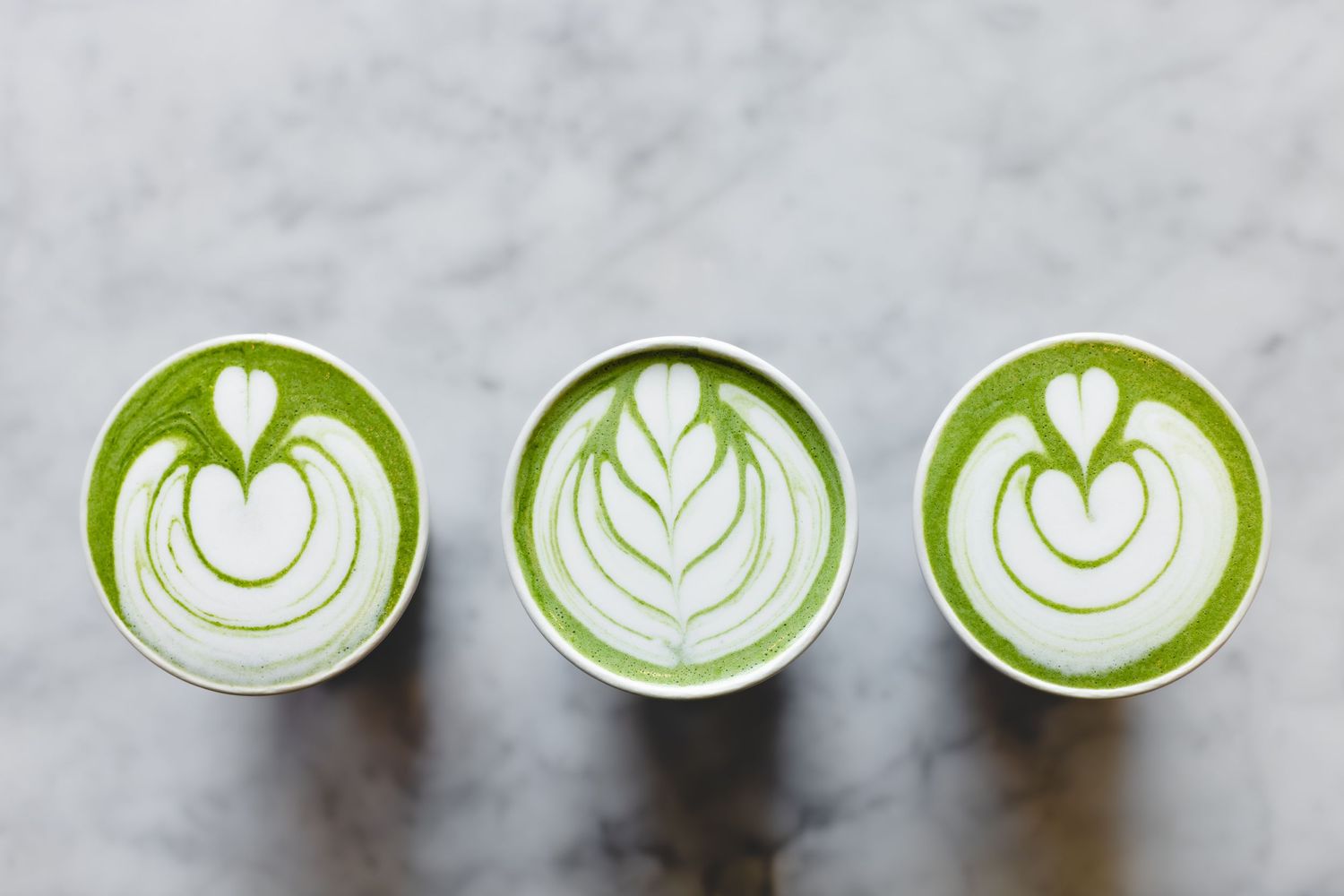 Three green matcha lattes with intricate white heart and leaf latte art designs on white marble surface.