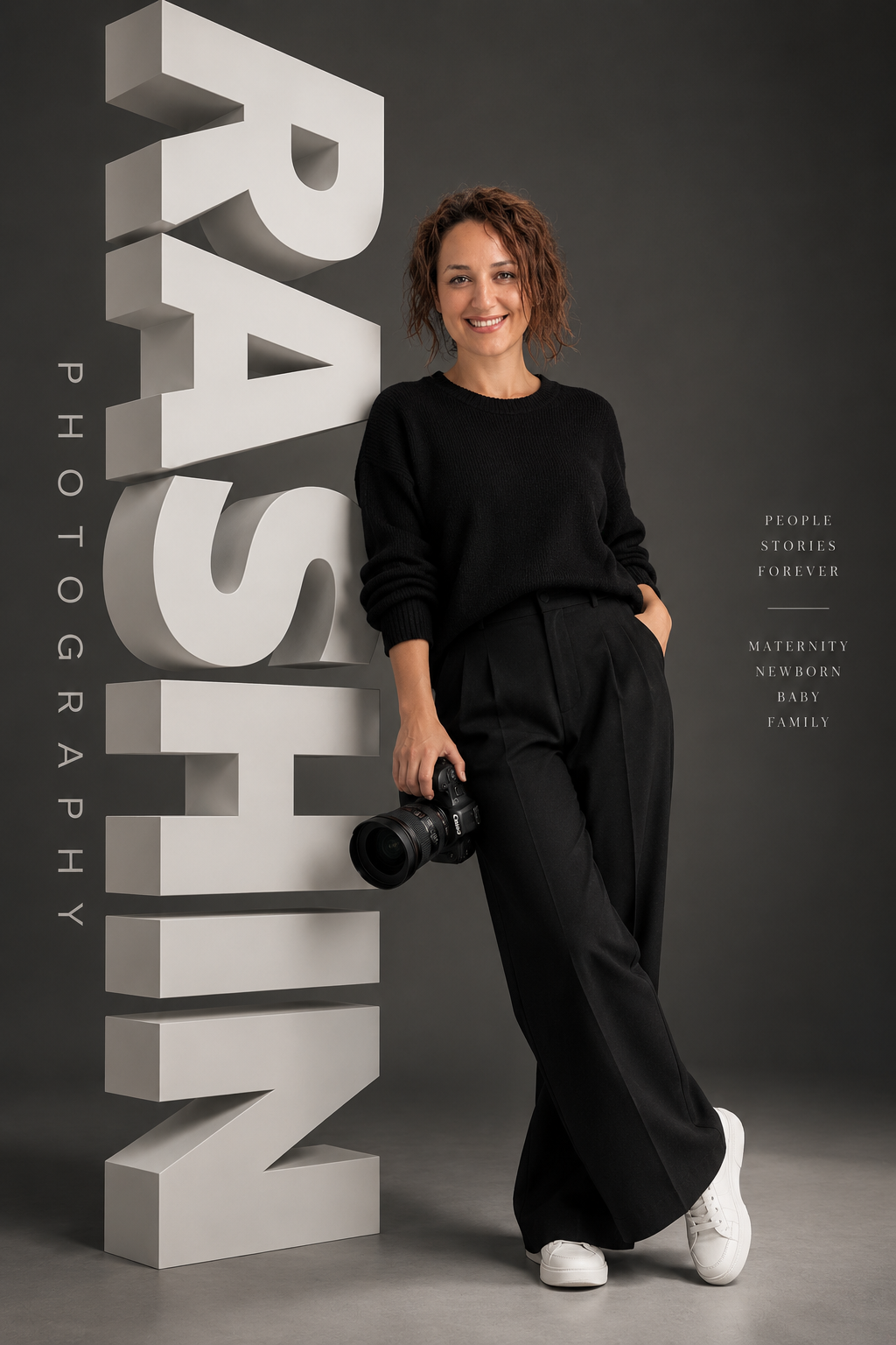 Professional photographer in black long sleeve shirt smiling while holding a DSLR camera against white backdrop.