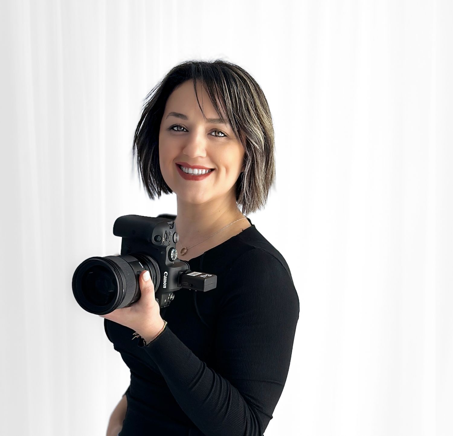 Professional photographer in black long sleeve shirt smiling while holding a DSLR camera against white backdrop.