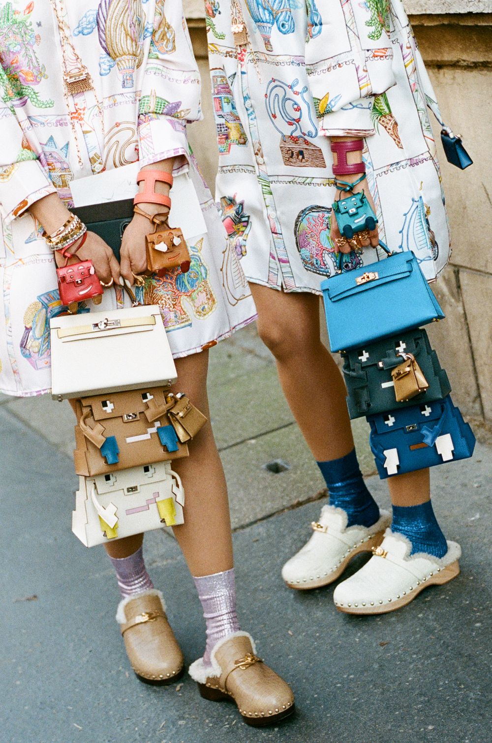Legs wearing colorful printed dresses, whimsical miniature handbags, socks and clogs on a street.
