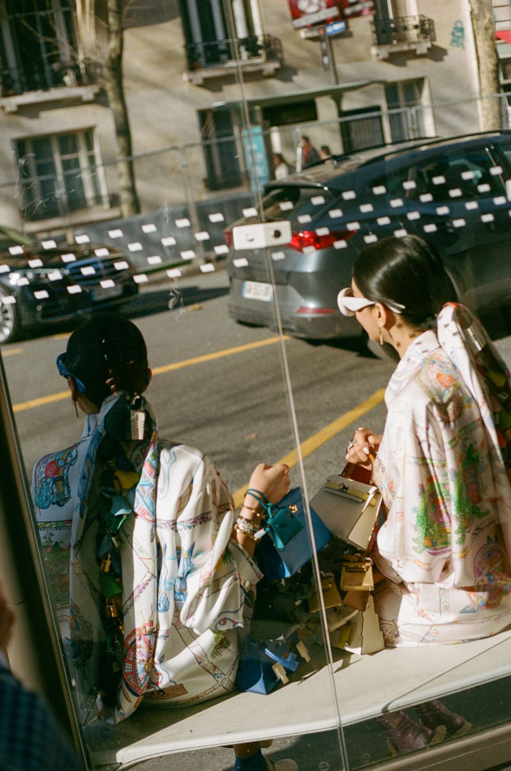 People wearing face masks sit together on steps along an urban street while viewed through reflective glass.