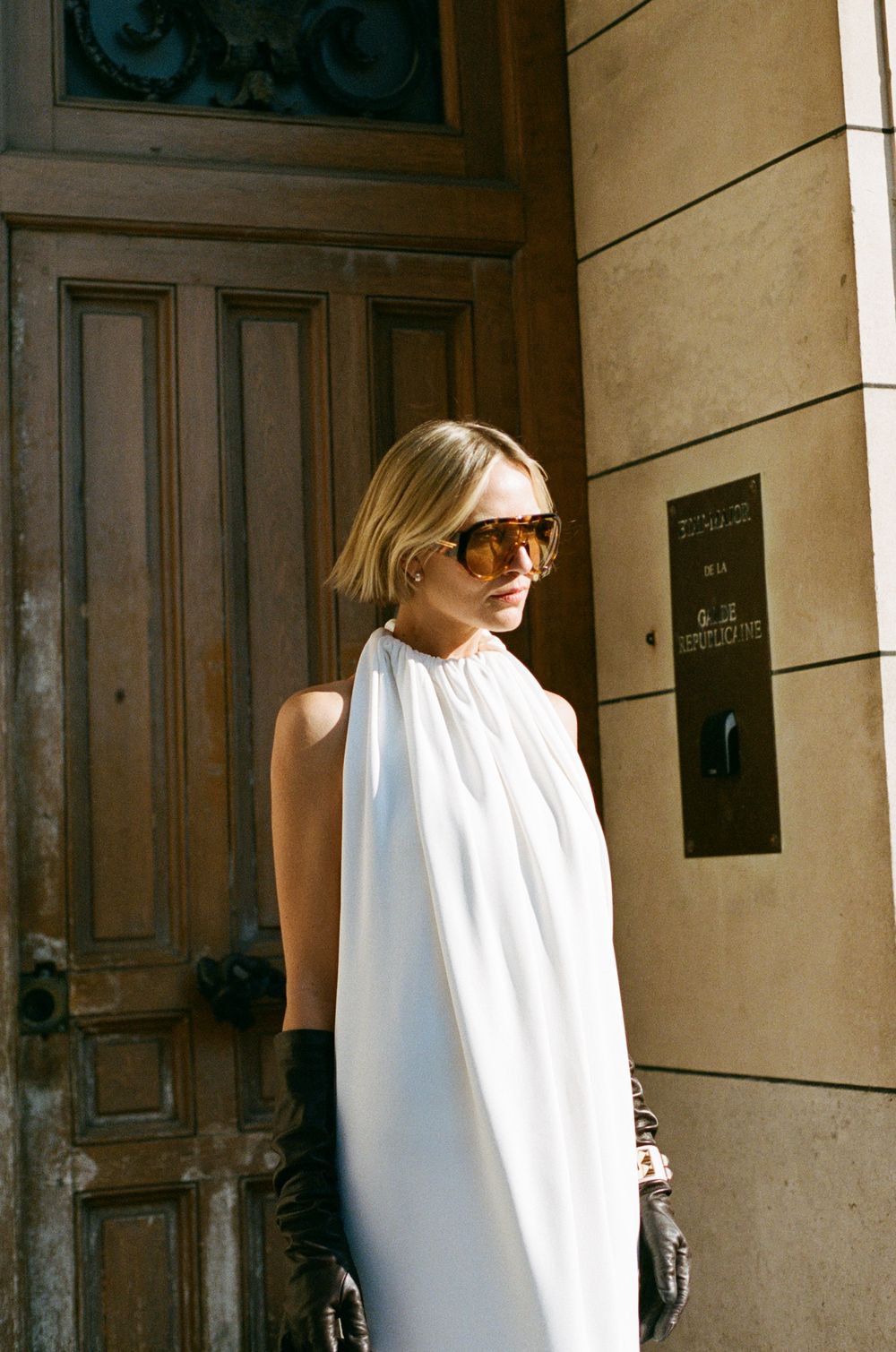A person in a sleek white high-neck dress and sunglasses stands elegantly against a wooden door in warm sunlight.
