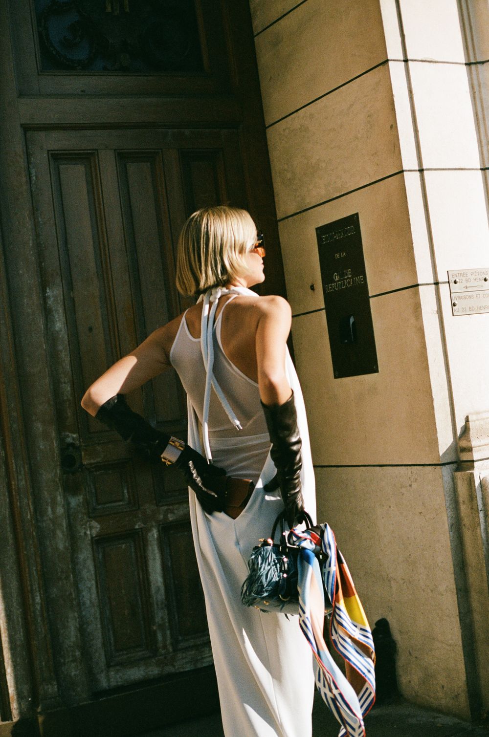 Figure in white sleeveless dress carrying bags stands outside vintage doorway in warm sunlit setting.