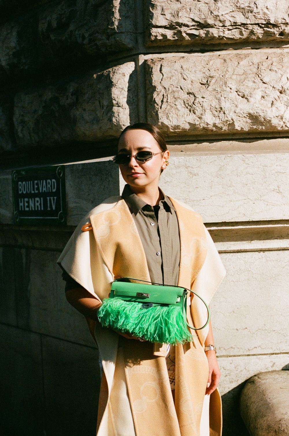Street fashion with beige coat and vibrant green feathered handbag against stone wall background.