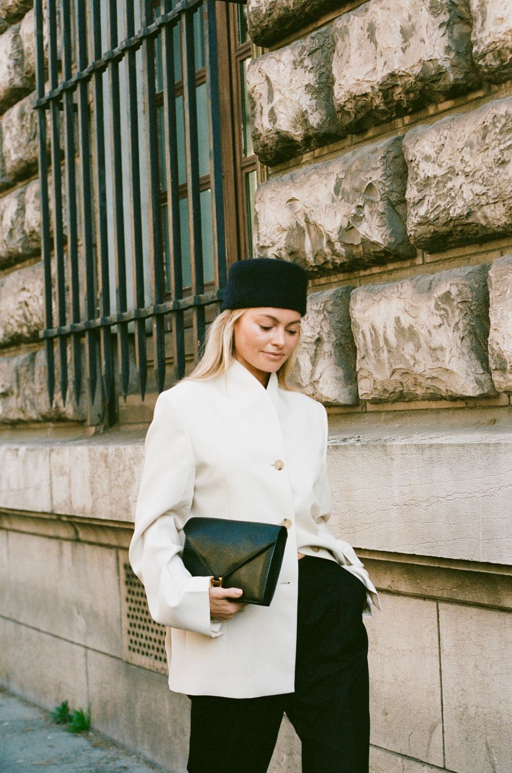 A stylish winter outfit with a white coat, black beret, and clutch bag photographed against a stone wall with iron bars.