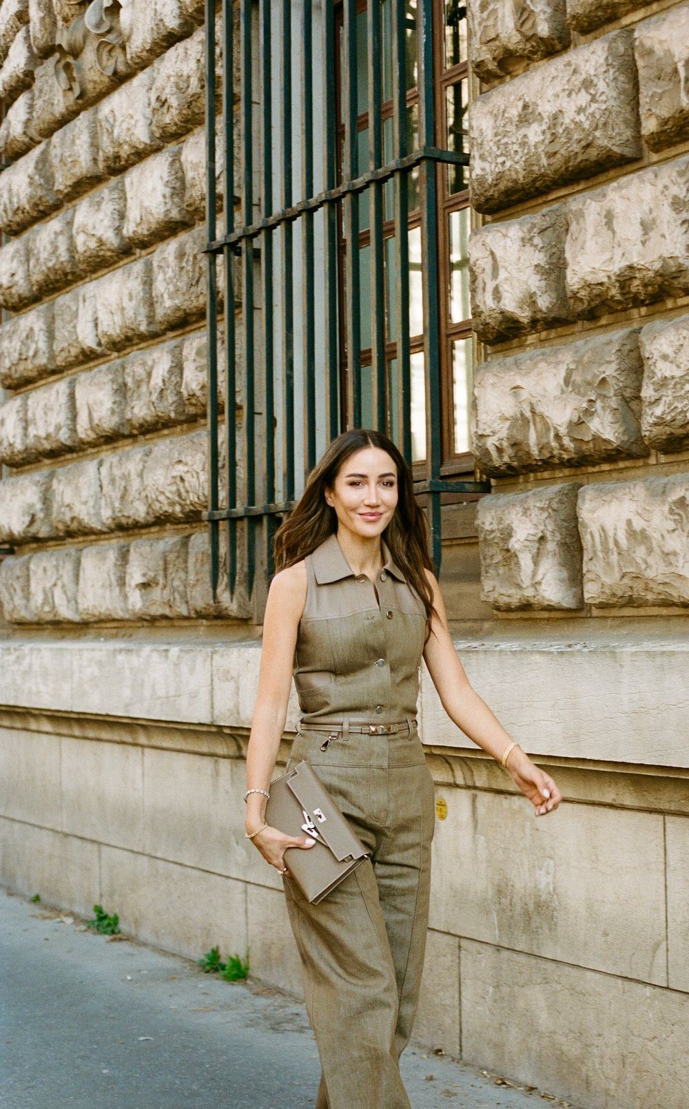 Woman wearing olive green jumpsuit stands against a stone wall with metal bars and holds a metallic clutch purse.