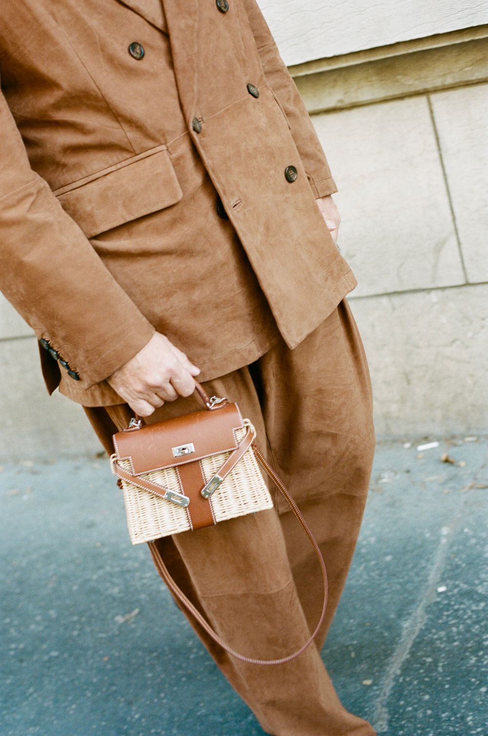Person wearing a brown tailored suit carries a cognac leather and woven straw handbag while walking on a sidewalk.