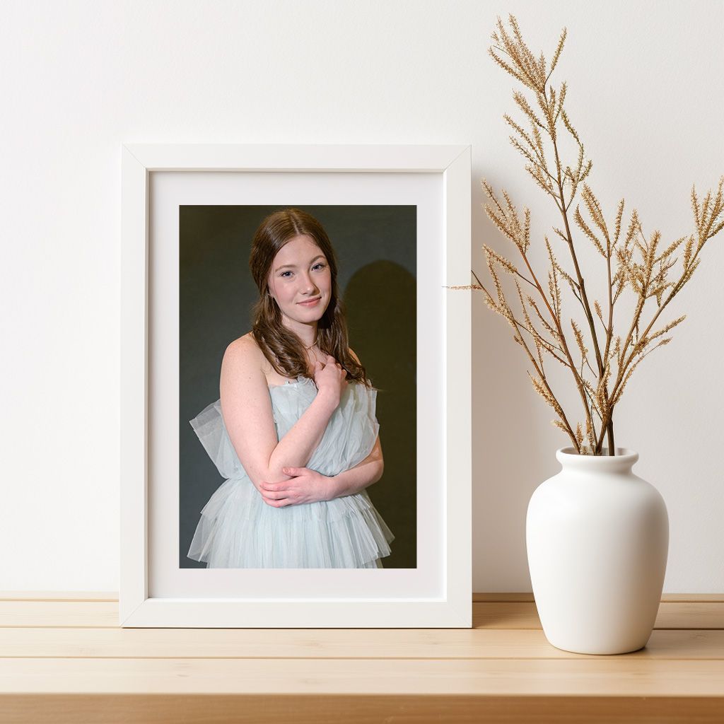 White framed portrait photograph displayed on wooden table with minimalist white vase and dried branches as decor.