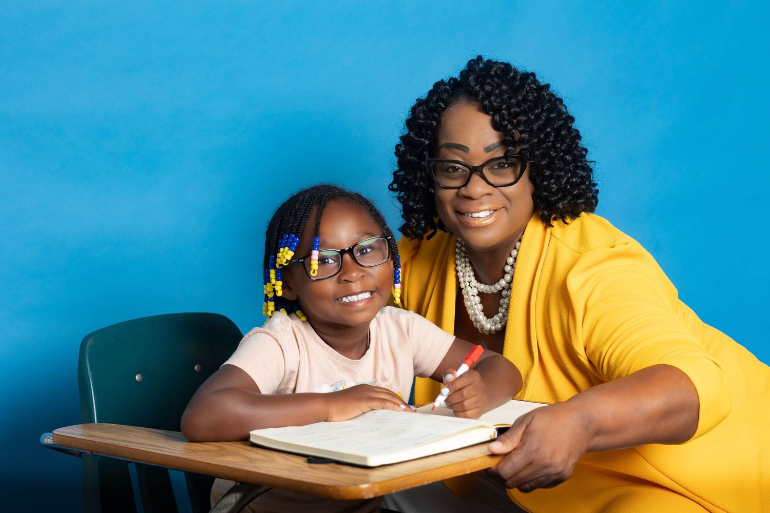 A teacher in a yellow cardigan helps a young girl with glasses with her schoolwork at a desk against a blue background.