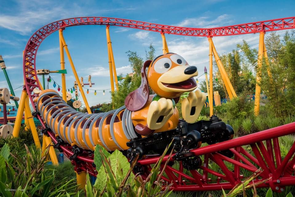 Slinky Dog rollercoaster track winds through Disney's Toy Story Land with vibrant red and yellow supports against blue sky.