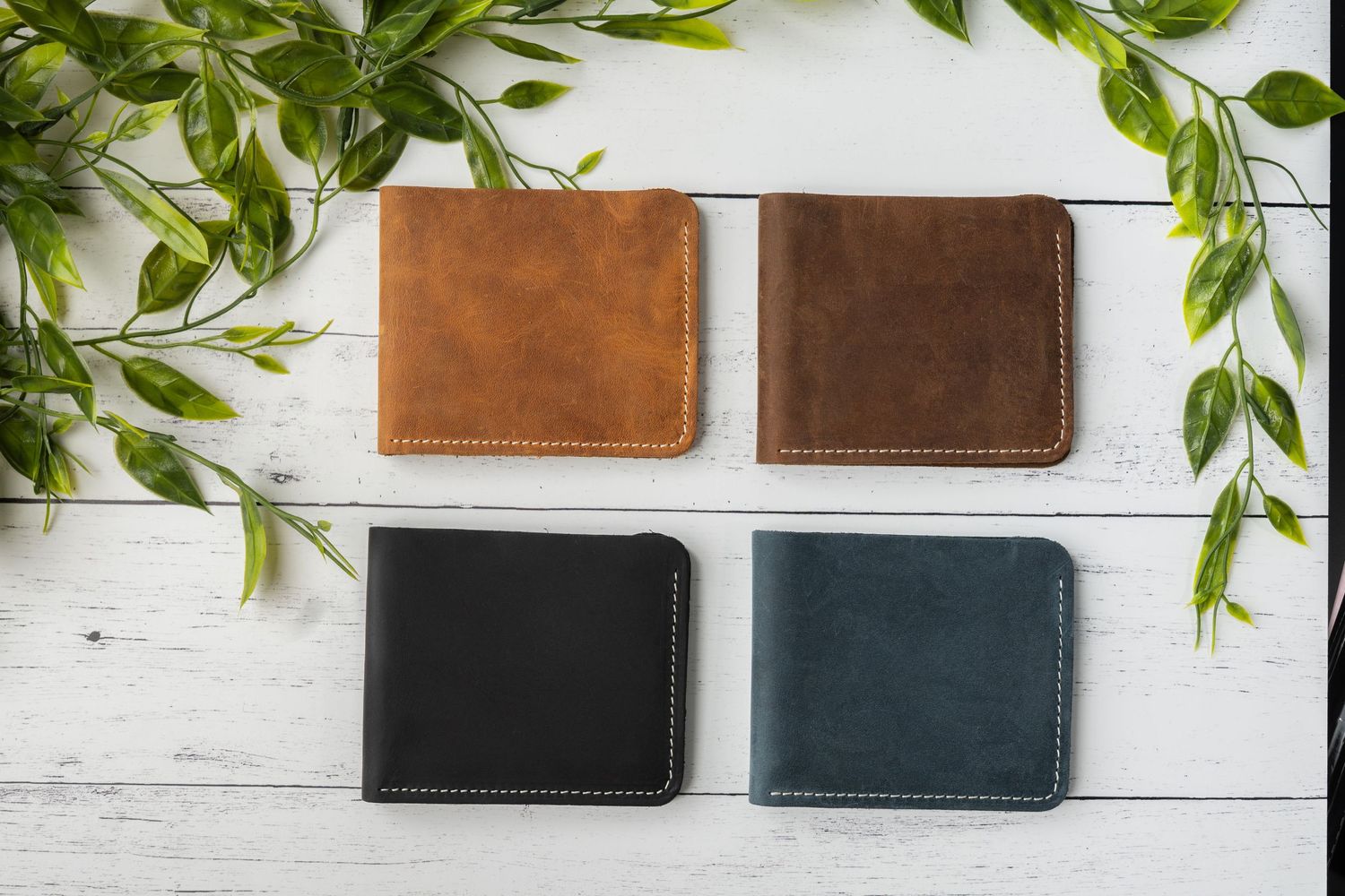Four leather wallets in tan, brown, black and blue arranged on white wooden surface with green foliage decoration.
