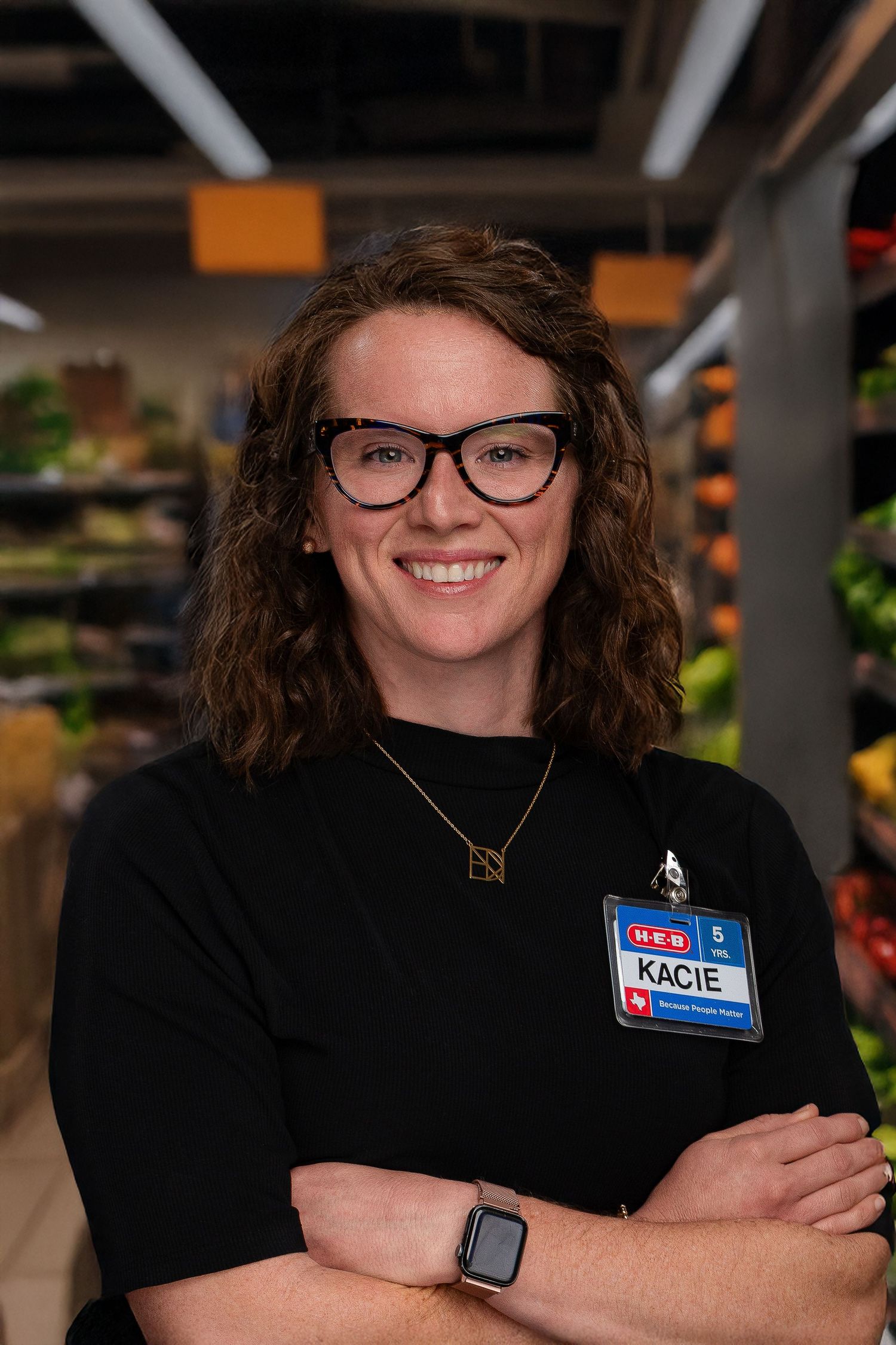 Retail employee in black uniform and glasses poses with arms crossed in grocery store setting.