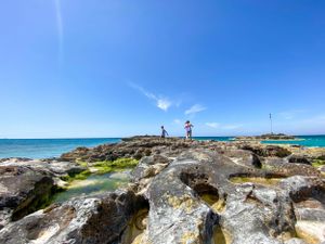 People explore rocky coastal formations along turquoise waters under bright blue skies in Batroun, Lebanon.