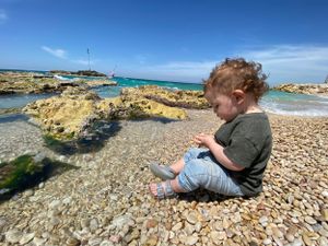 A young child sits on a rocky coastline overlooking bright blue waters and rugged cliffs on a sunny day.