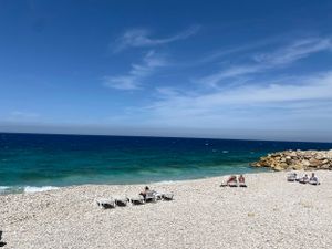 Turquoise Mediterranean waters meet a pristine pebble beach with lounge chairs under bright blue skies in Batroun, Lebanon.