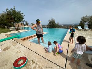 Friends and family gather around a scenic infinity pool overlooking a landscape on a sunny day.