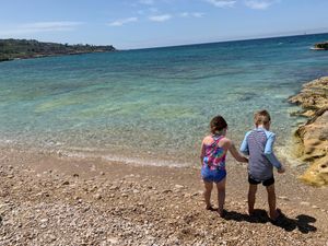 Two children explore a rocky Mediterranean beach with turquoise waters and clear blue skies in the background.