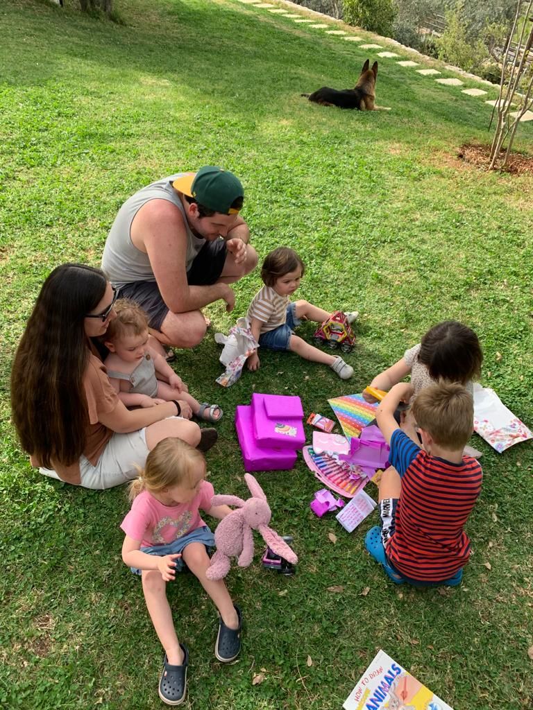 A group sits on a lawn having a picnic with toys and books while a dog rests in the background on a sunny day.