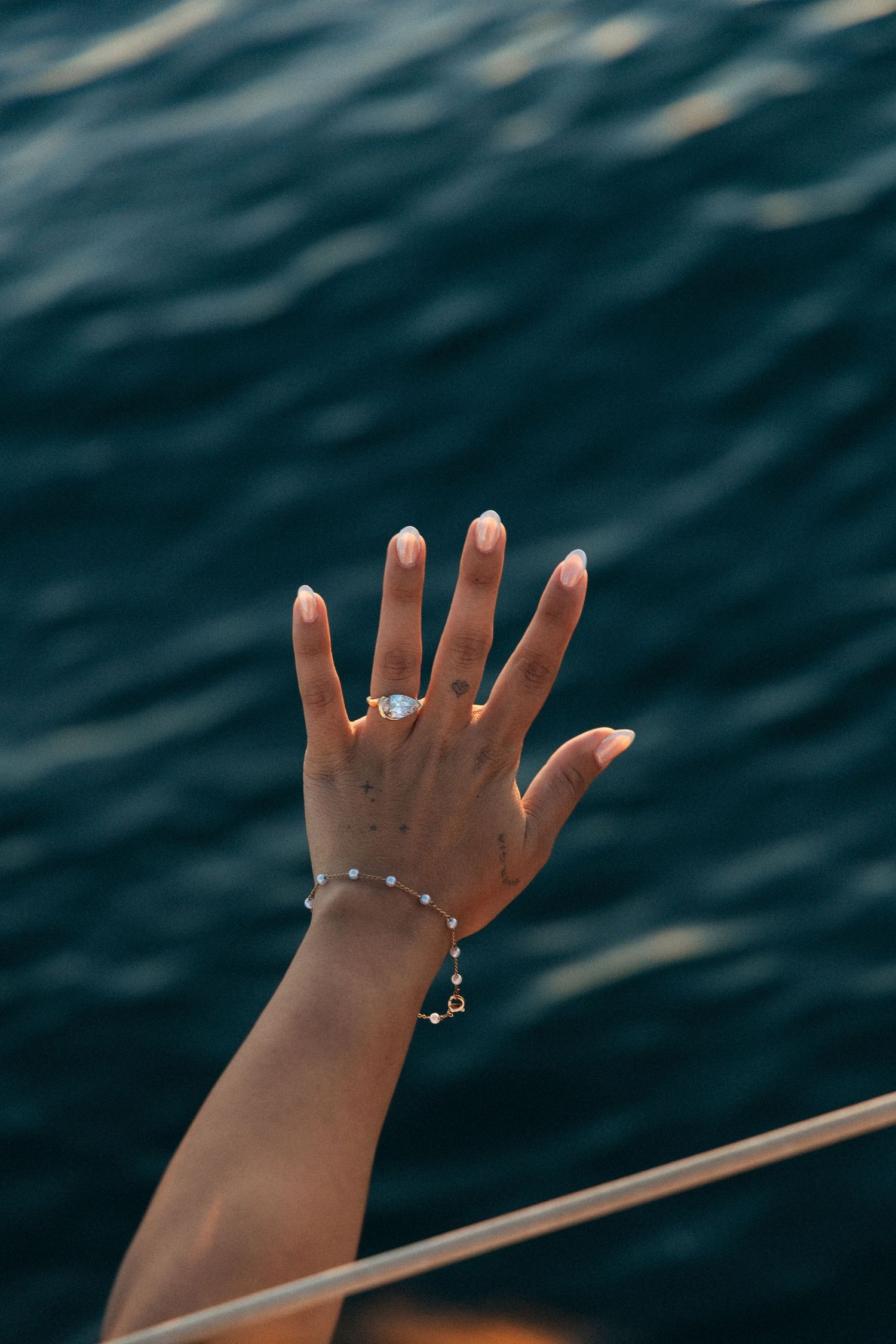 A feminine hand with an engagement ring and delicate bracelet reaches up against a backdrop of dark rippling water.